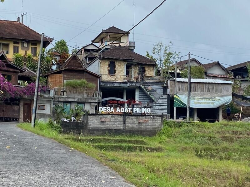Papan nama Desa Adat Piling di tepi jalan desa dengan latar lahan hijau